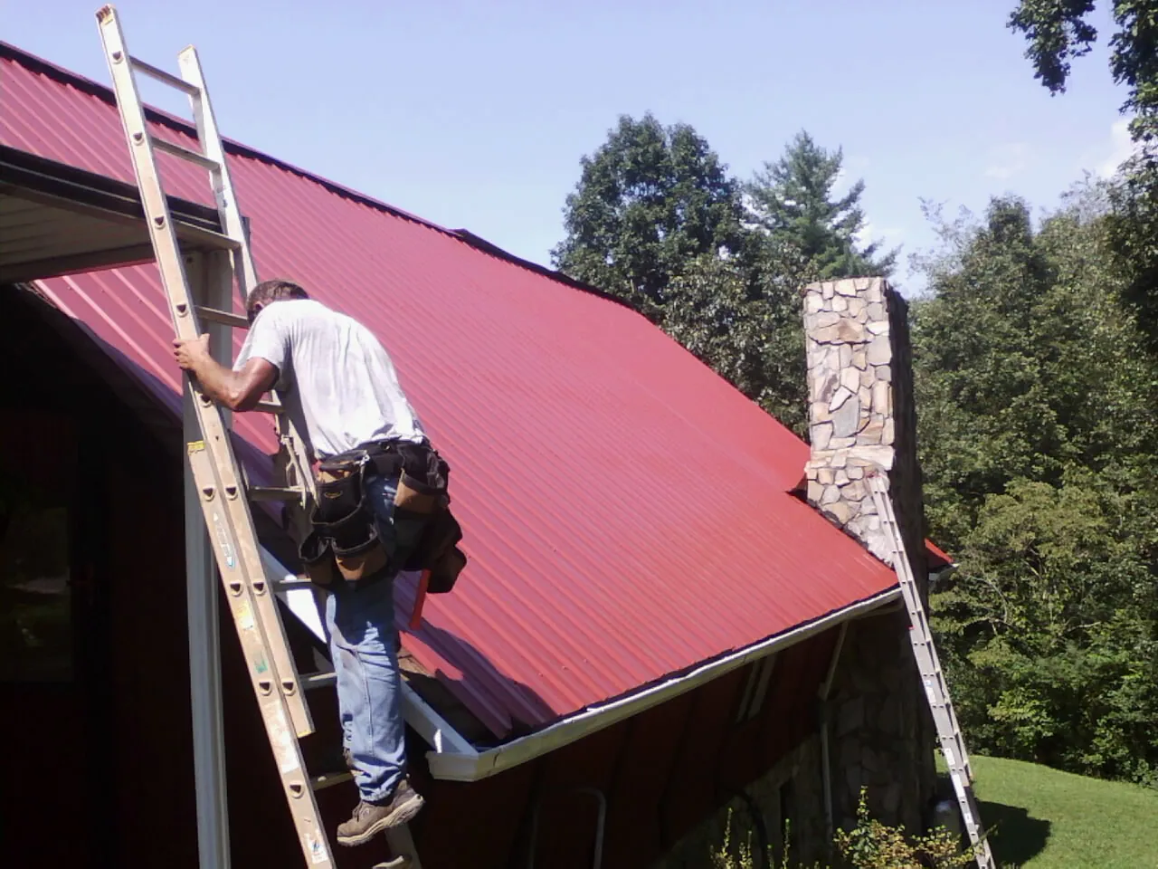 Roofer on a ladder beside a red metal roof and stone chimney — Soffit & Fascia Repair by Roofing Brothers in Kindred
